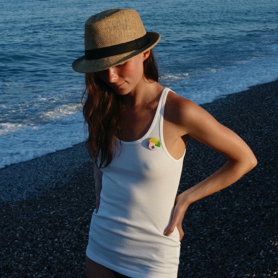 A woman wearing a straw fedora with a black band and a white tank top with a small colorful pin on the chest, standing on a dark pebble beach with the ocean in the background.