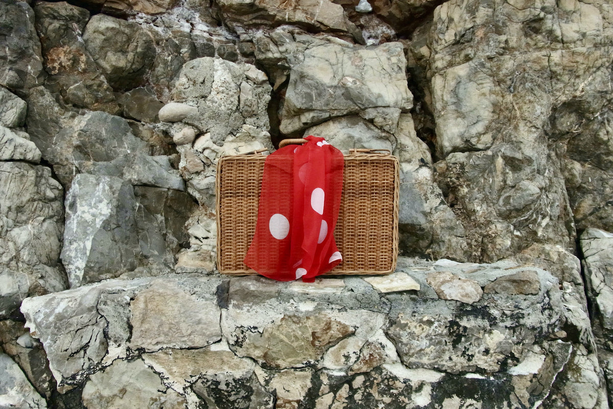 A brown wicker picnic basket with a red scarf featuring white polka dots, set against a rugged gray rock wall.