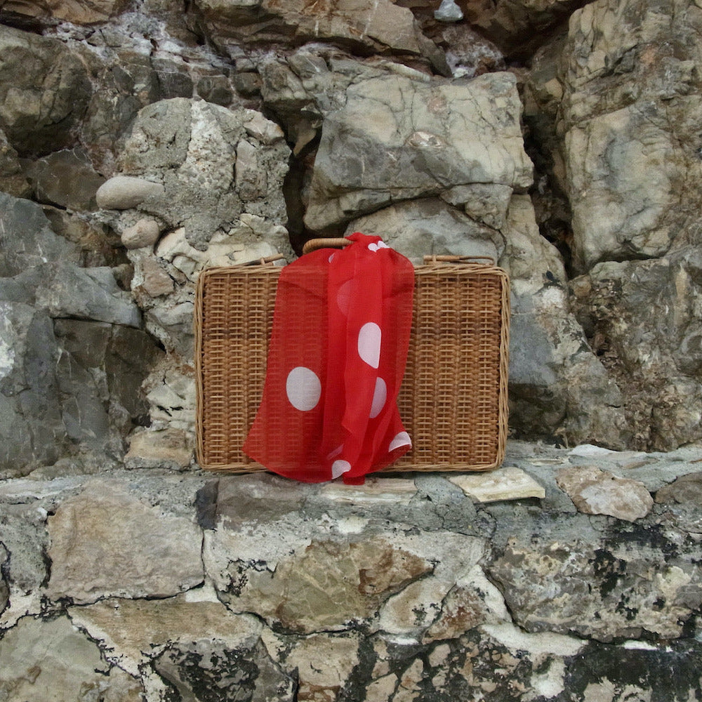 A brown wicker picnic basket with a red scarf featuring white polka dots, set against a rugged gray rock wall.